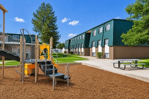 A playground with a yellow slide and a green building in the background at The Morgan Apartments, Layton