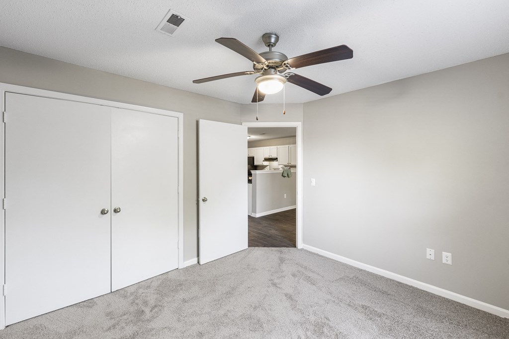 Modern apartment bedroom with light gray walls, plush gray carpeting, and a ceiling fan with a built-in light fixture. A white double-door closet is on one side, and an open door leads to a kitchen with white cabinetry and dark wood-style flooring