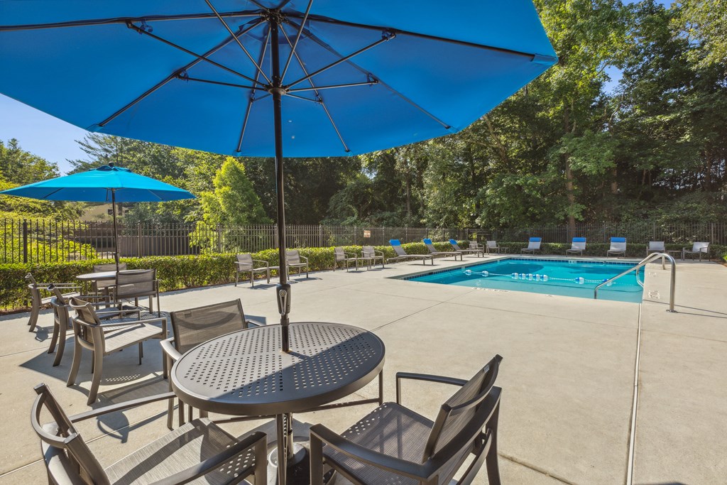 A blue umbrella shades a table and chairs by a pool.