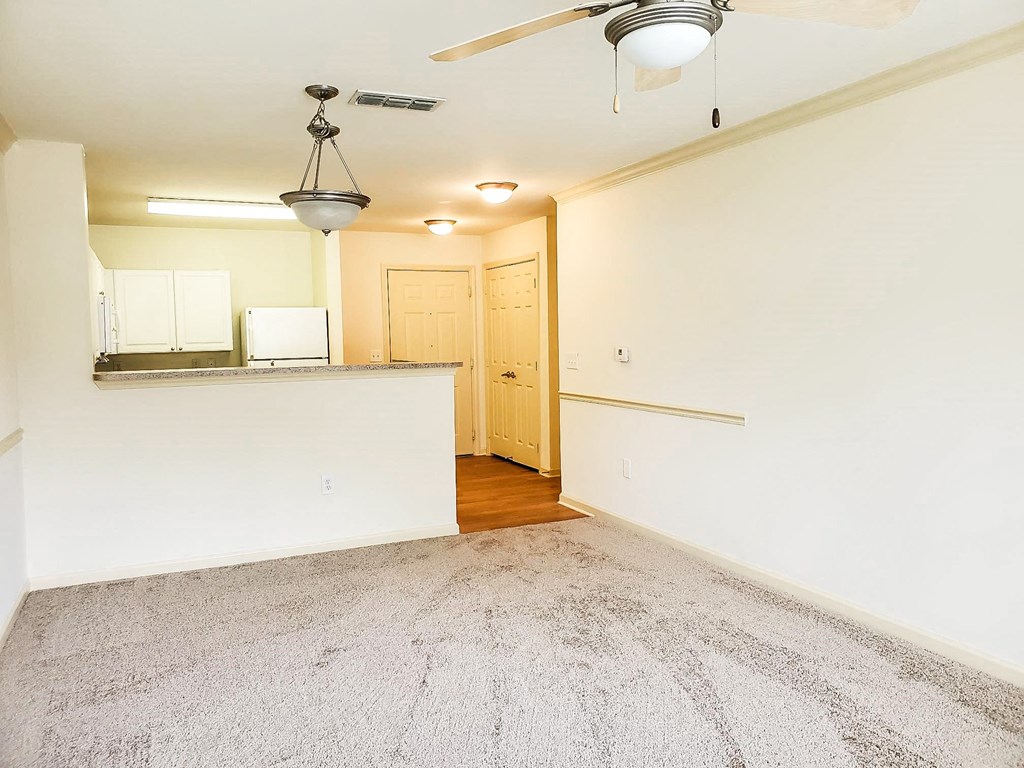 Empty apartment with a carpeted living area, an open kitchen with white cabinets and countertops, and a breakfast bar. A ceiling fan and pendant light hang from the ceiling. The entrance and closet doors are visible in the background with wood flooring