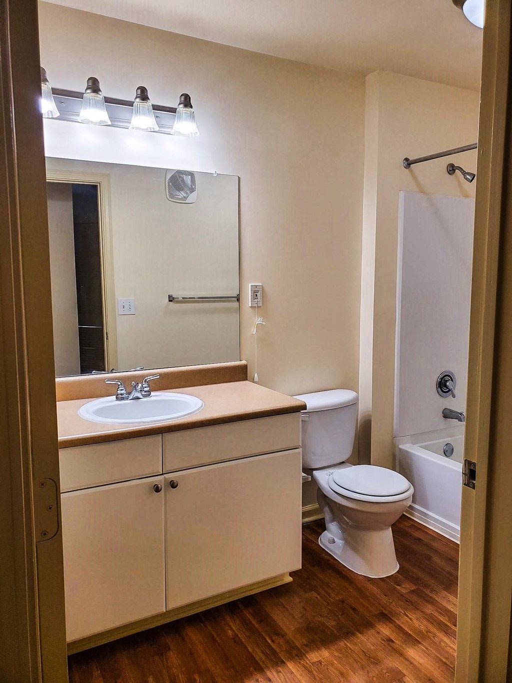 A bathroom with a white vanity, beige countertop, and a large mirror with overhead lighting. It features a toilet, a bathtub with a shower rod, and wood-style flooring