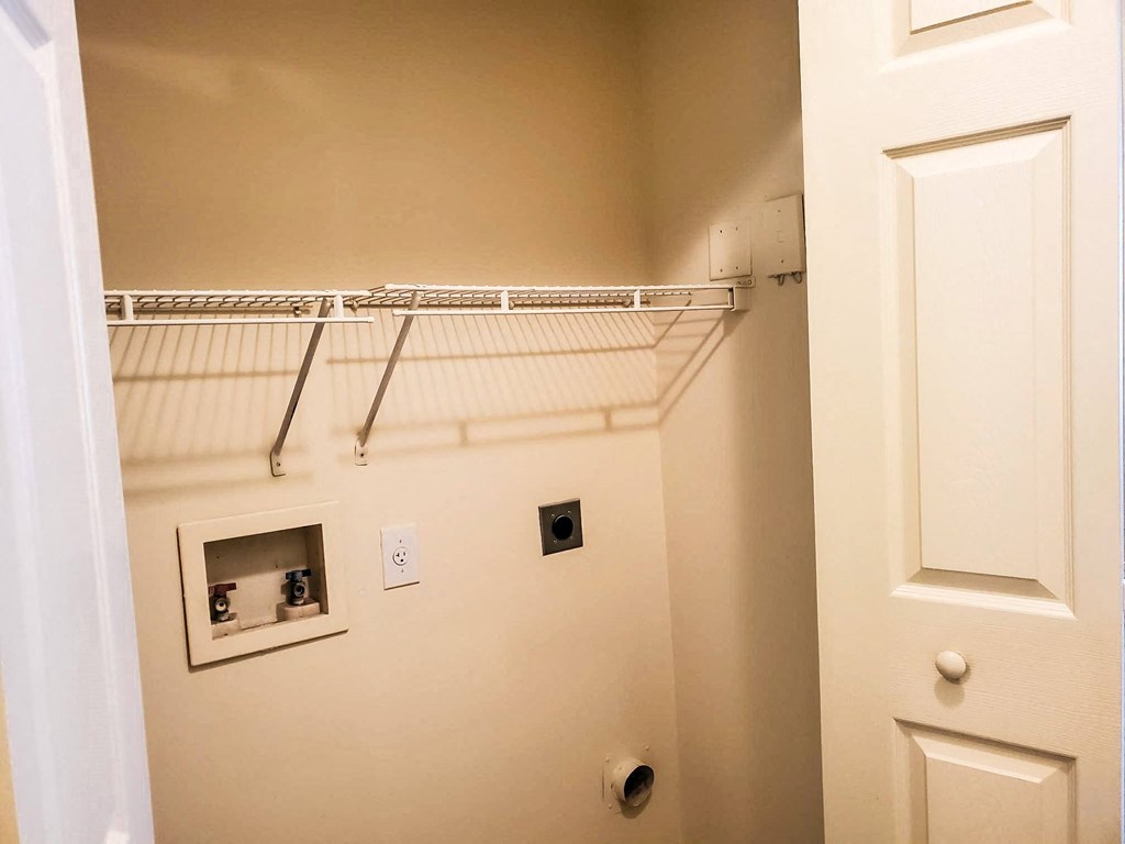 A small laundry area with beige walls, a white wire shelf, and hookups for a washer and dryer. A partially open white bifold door is visible on the right side
