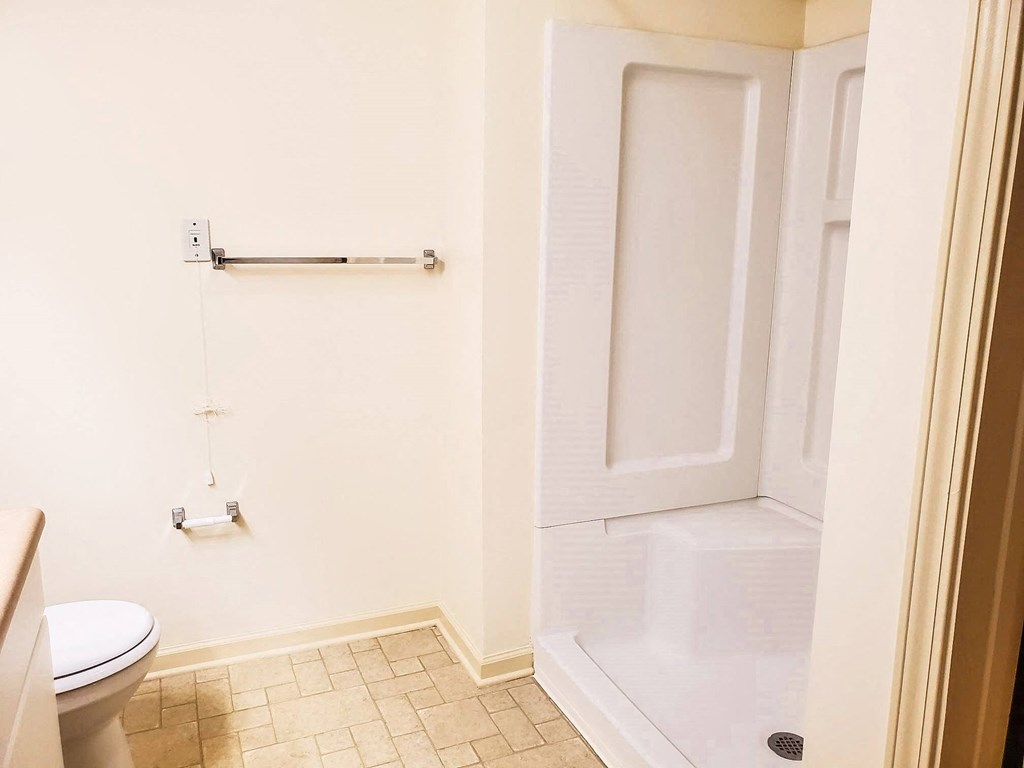 A bathroom with a white walk-in shower that has built-in seating, a toilet, and a beige countertop. The floor has light-colored tile, and a towel rack and toilet paper holder are mounted on the wall