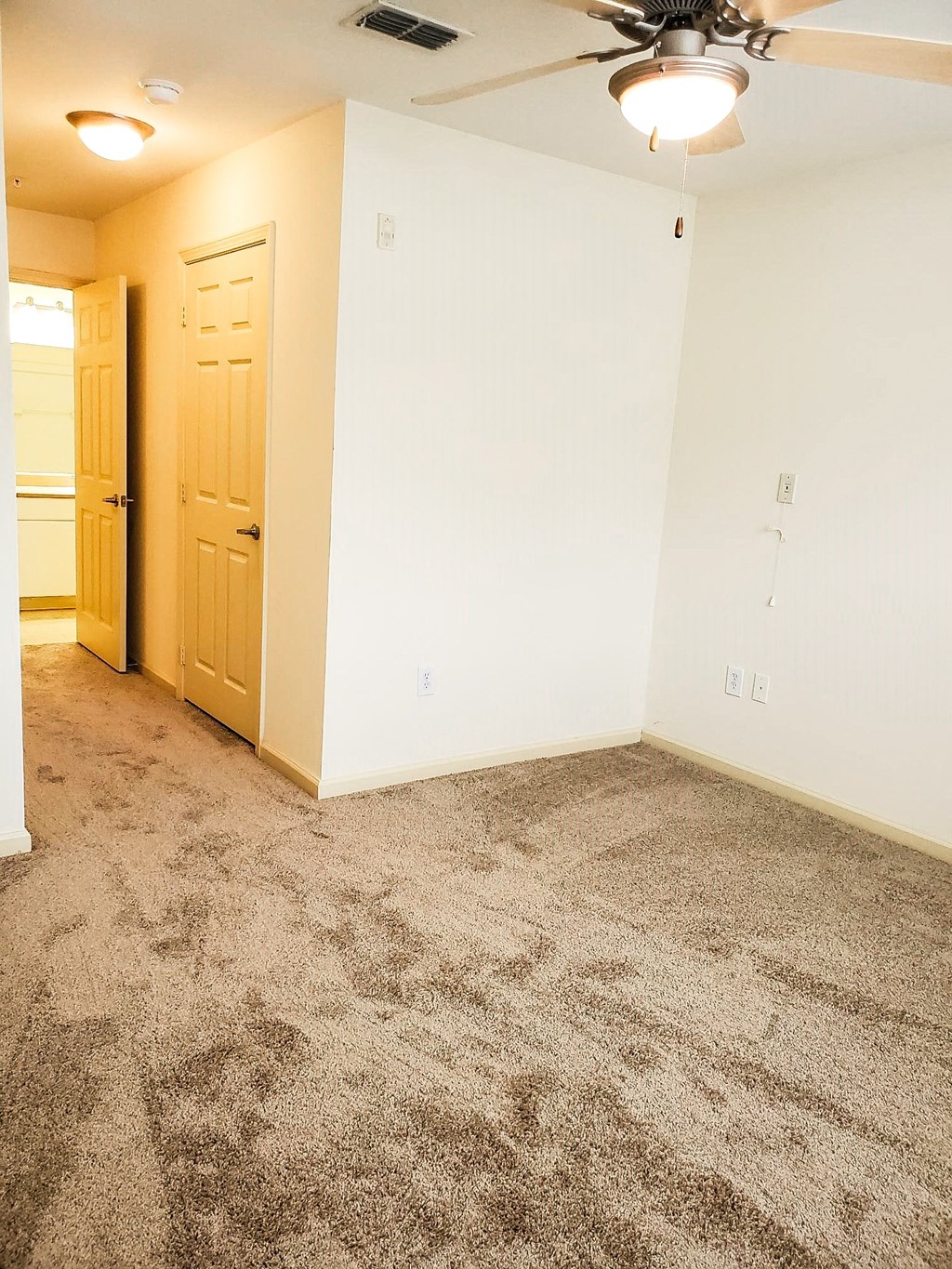 An empty bedroom with beige carpeting, off-white walls, and a ceiling fan with a light. A partially open door leads to a hallway and a laundry area is visible in the background