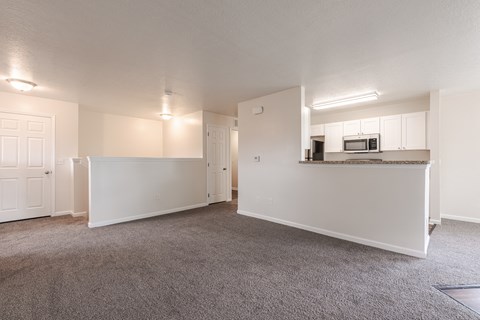 the living room and kitchen of an apartment with carpeted flooring and white cabinets
