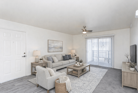 A living room with a grey couch, a white door, a ceiling fan, and a television.
