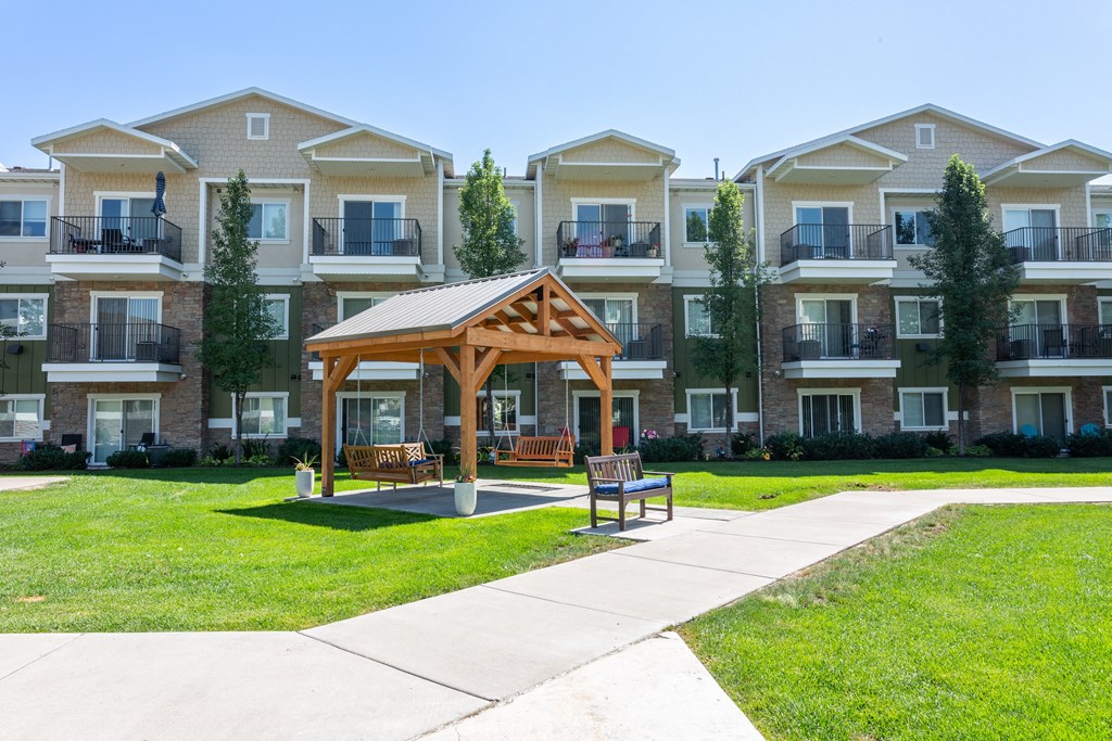 A wooden pavilion is in the middle of a grassy area in front of apartment buildings.
