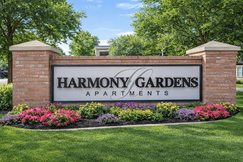 A sign for Harmony Gardens Apartments is framed by flowers and topped by two pillars.