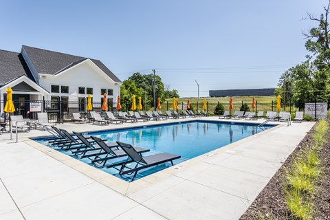 A pool with chairs and umbrellas is surrounded by a white building and a fence.