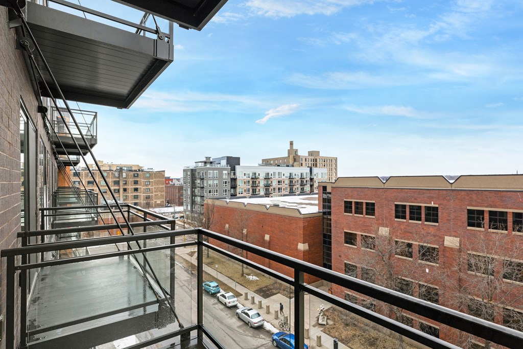 A balcony with a metal railing overlooks a parking lot and buildings.