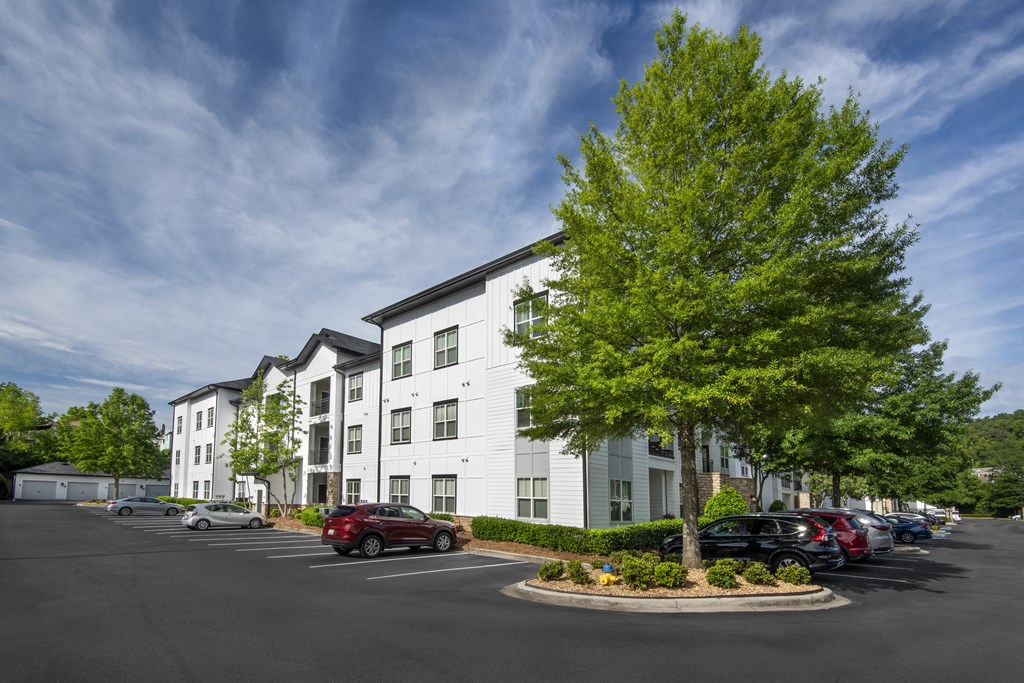 A parking lot in front of a white apartment building with a tree in the foreground.