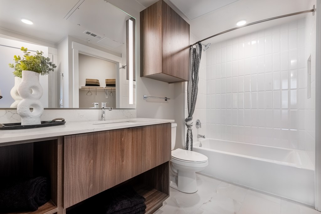 A white toilet sits in a modern bathroom with a wooden vanity and a white bathtub.