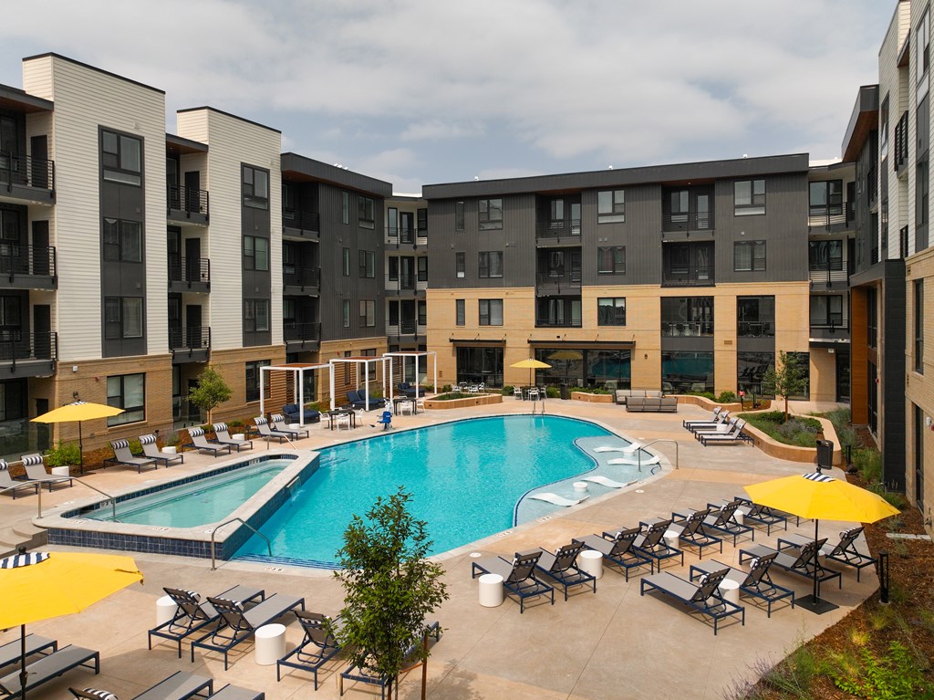 A large swimming pool surrounded by lounge chairs and umbrellas in front of apartment buildings.