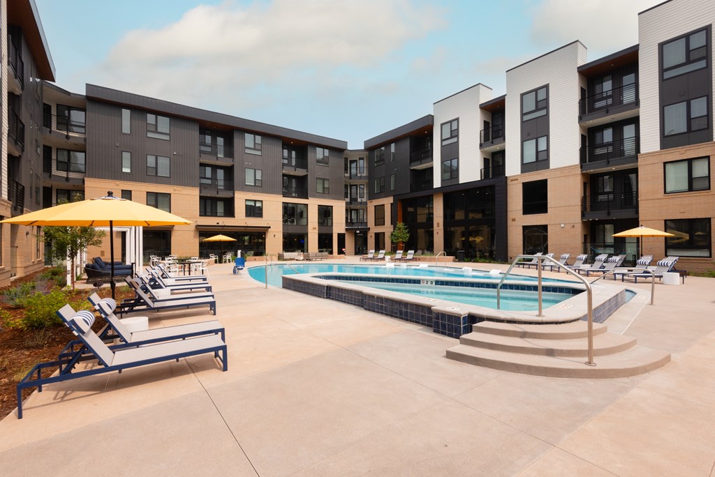 A swimming pool surrounded by sun loungers and umbrellas in front of apartment buildings.
