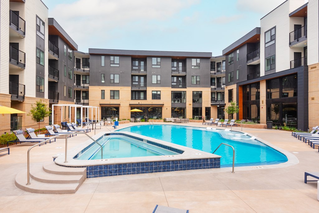 A large swimming pool is surrounded by lounge chairs in front of apartment buildings.