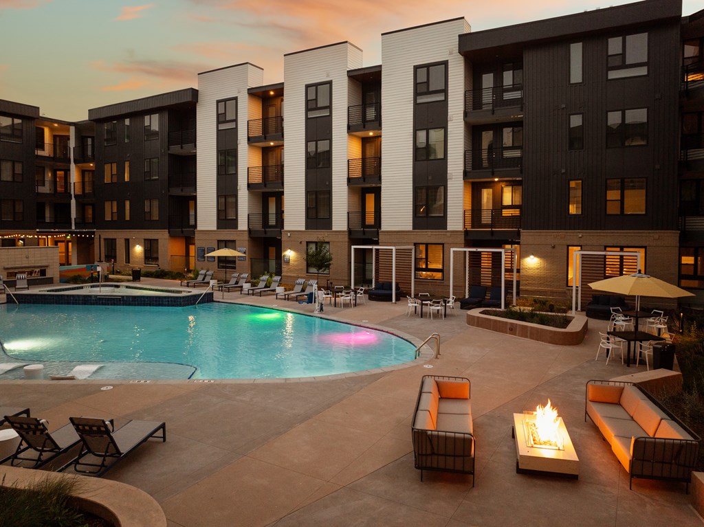 A large outdoor pool area with lounge chairs and a fire pit in front of apartment buildings.