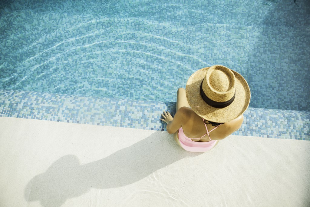 A woman in a straw hat and pink bikini sits by a pool.