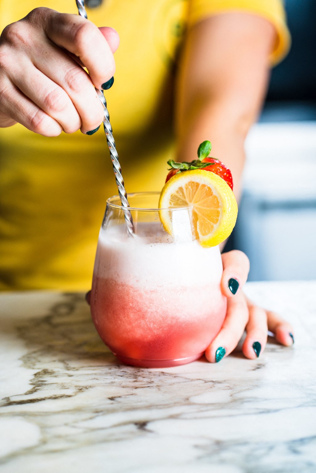 A person in a yellow shirt is stirring a pink drink with a lemon slice and a strawberry on the rim of the glass.
