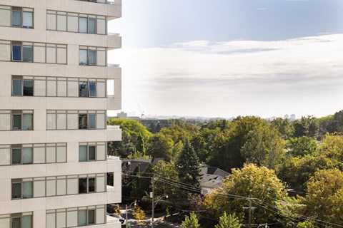 A tall white building with many windows is in the foreground with trees and houses in the background.