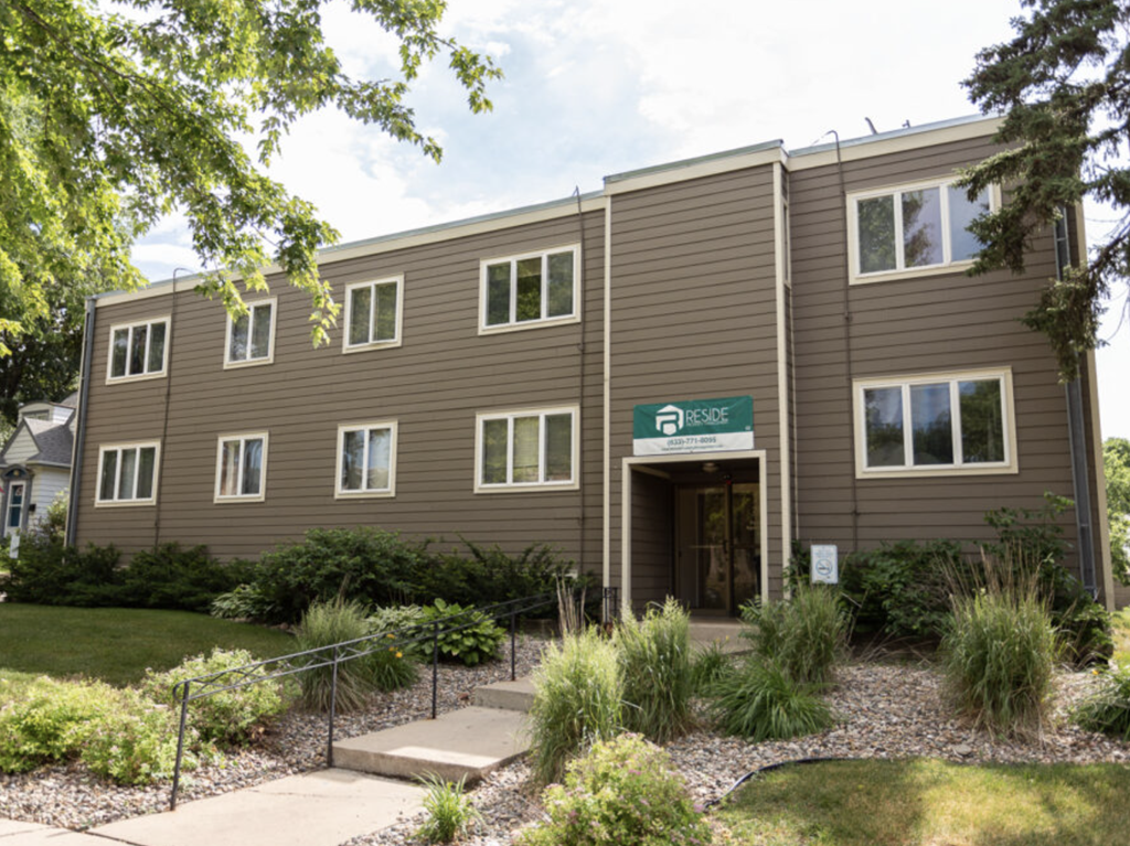 the front of a brown building with a sidewalk and plants