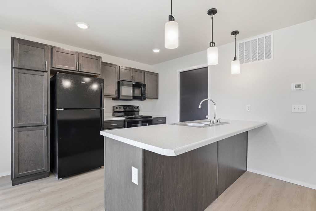 A modern kitchen with a black refrigerator and microwave, a white countertop, and wooden cabinets.