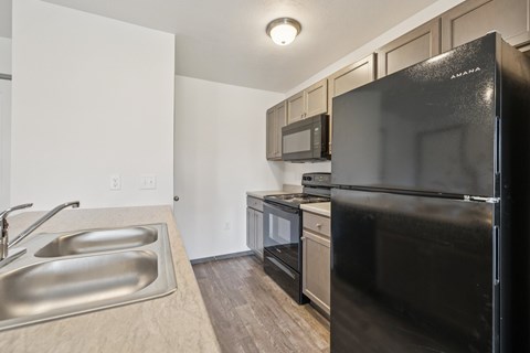 A black refrigerator is in the corner of a kitchen with a sink and microwave above it.