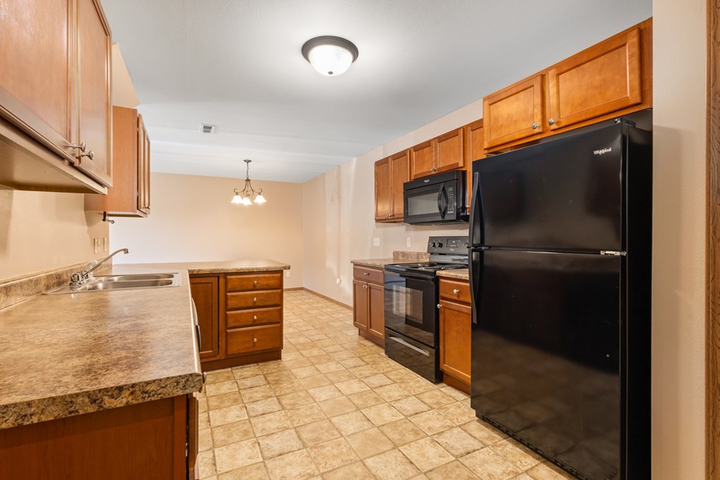 A kitchen with a black refrigerator, brown cabinets, and a granite countertop.