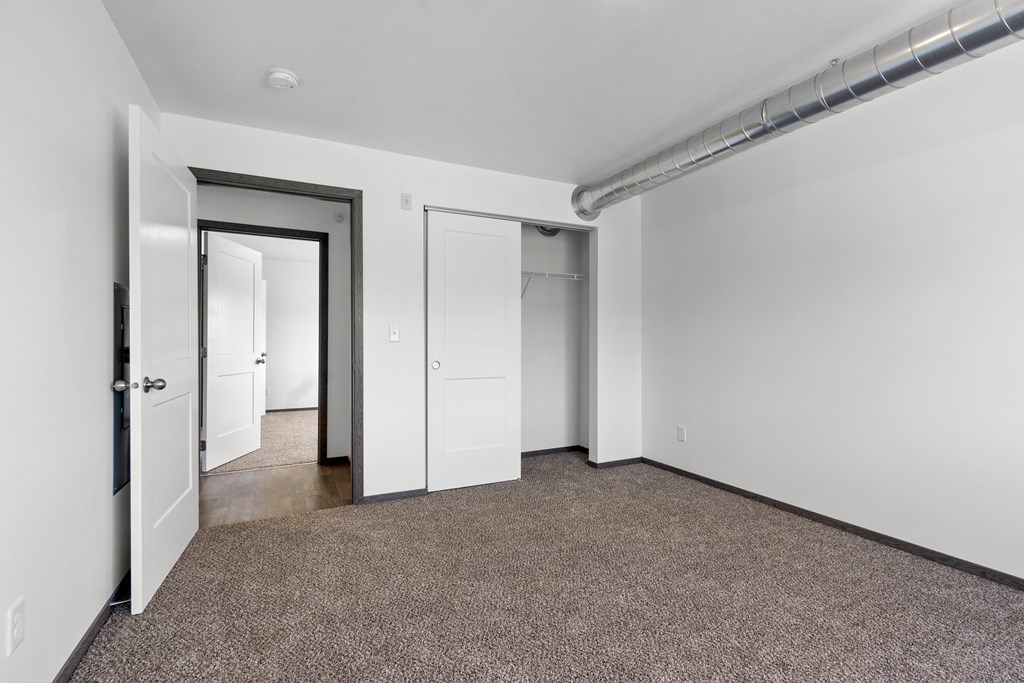 A room with a carpeted floor and white walls, with a door on the left and a window on the right.