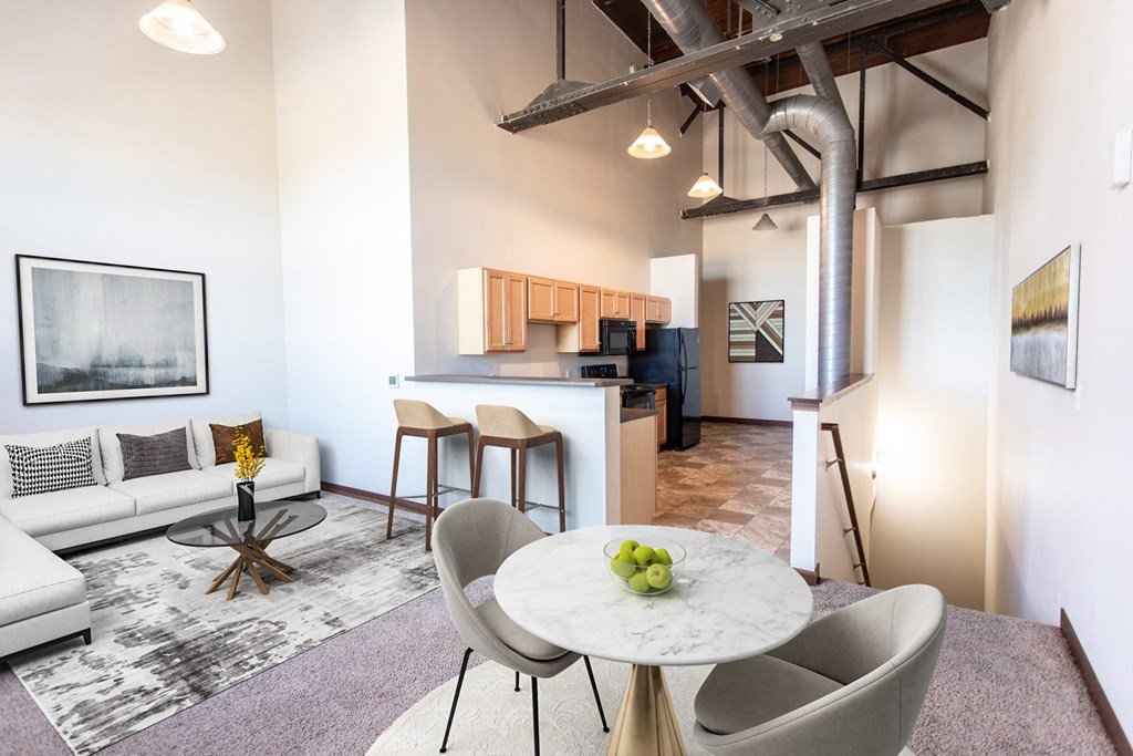 A modern living room with a white couch, a round table with a bowl of fruit on it, and a kitchen area in the background.