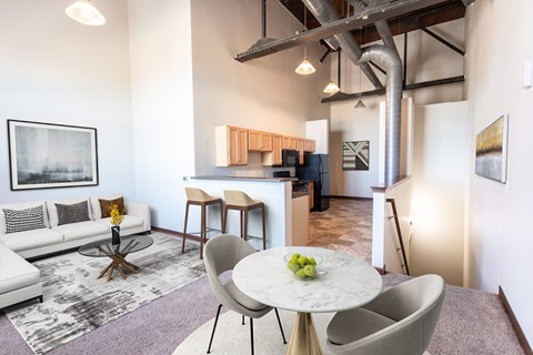 A modern living room with a white couch, a round table with a bowl of fruit on it, and a kitchen area in the background.