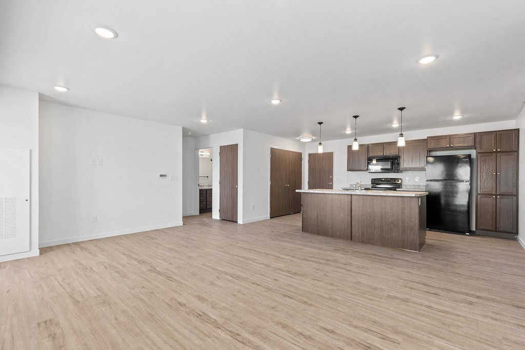 an empty living room with a kitchen with a refrigerator and a sink