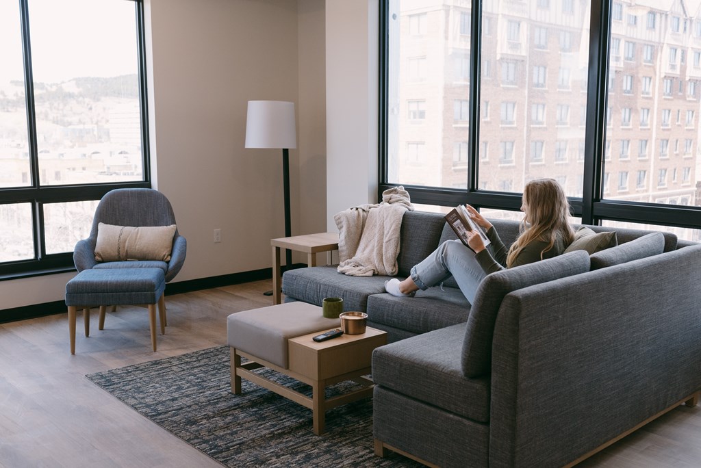 A woman is sitting on a grey couch in a living room with a large window behind her.