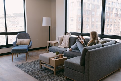 A woman is sitting on a grey couch in a living room with a large window behind her.