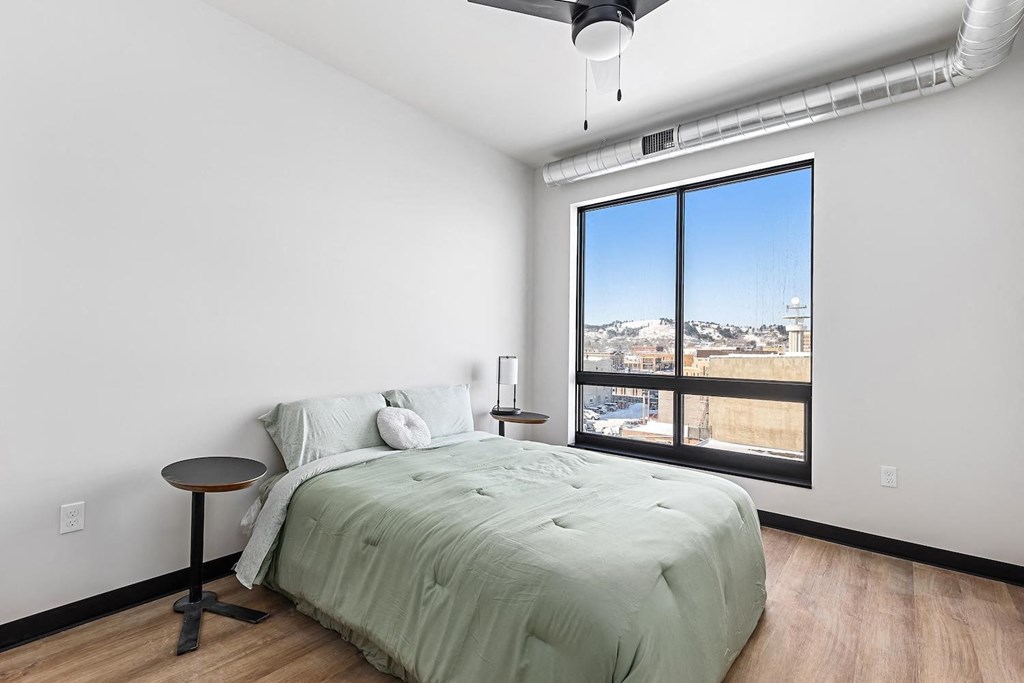 A bedroom with a bed, a small table, and a window overlooking a cityscape.
