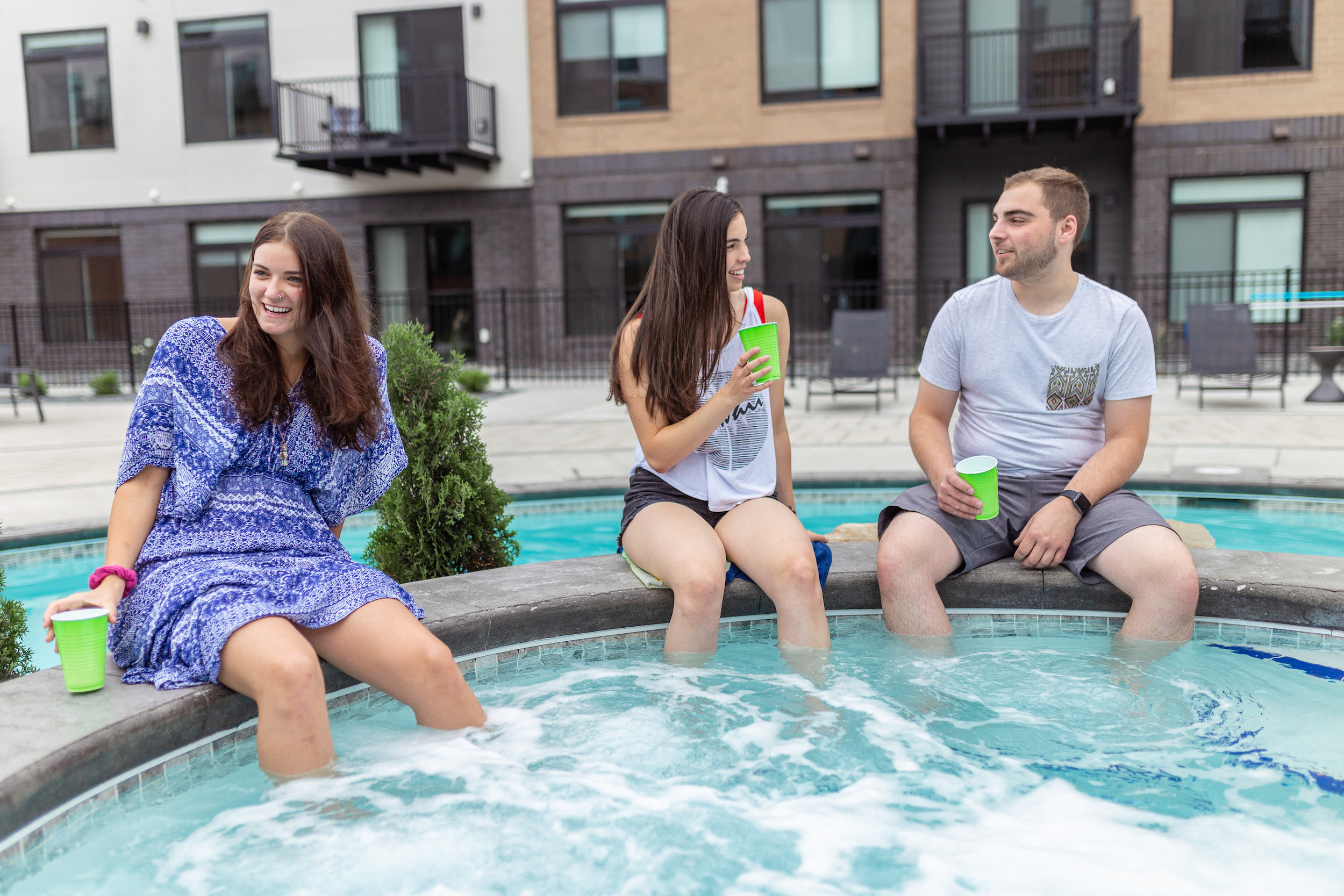 two women and a man sitting on the edge of a swimming pool with drinks