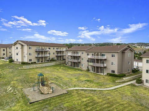 A playground with a slide is in the middle of a grassy area in front of apartment buildings.