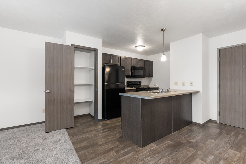 A kitchen with a black fridge and microwave, brown cabinets, and a grey rug.
