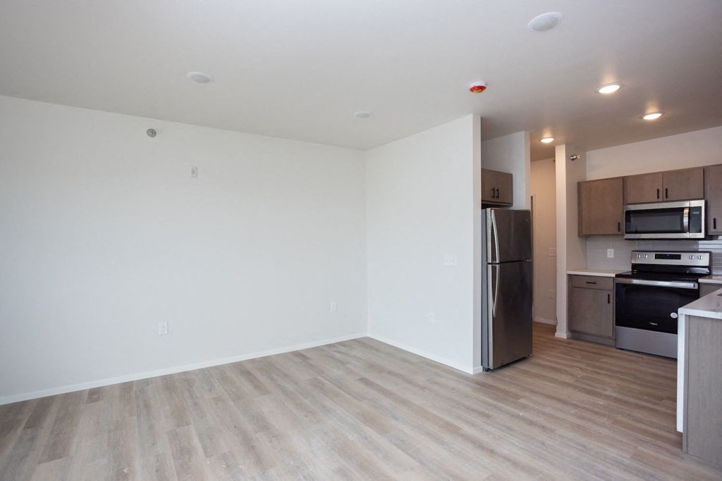 an empty living room with a kitchen with stainless steel appliances and wooden floors
