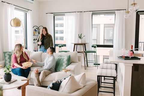 A woman is standing in a living room with a man sitting on a couch and a woman sitting on a chair.