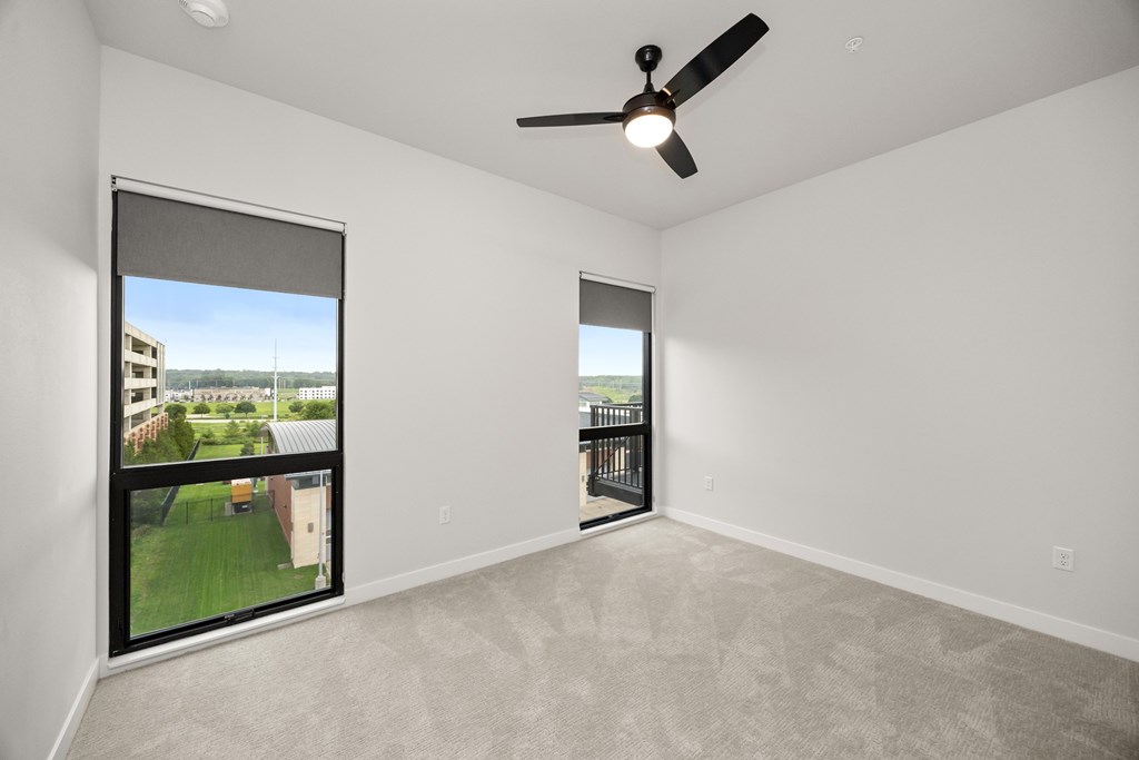 A room with a ceiling fan and a large window with a view of a building and a lawn.