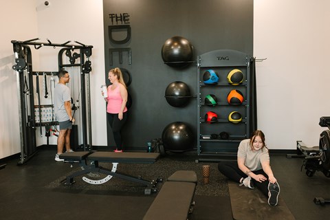 A woman is sitting on a mat in a gym, stretching her legs.