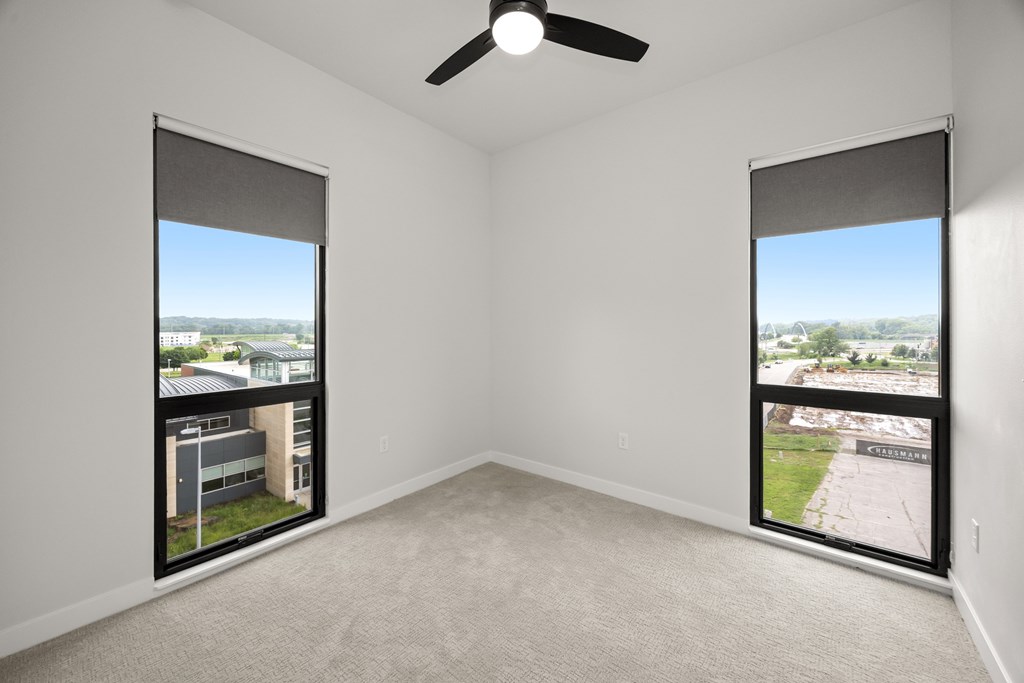A room with a ceiling fan and two windows overlooking a cityscape.