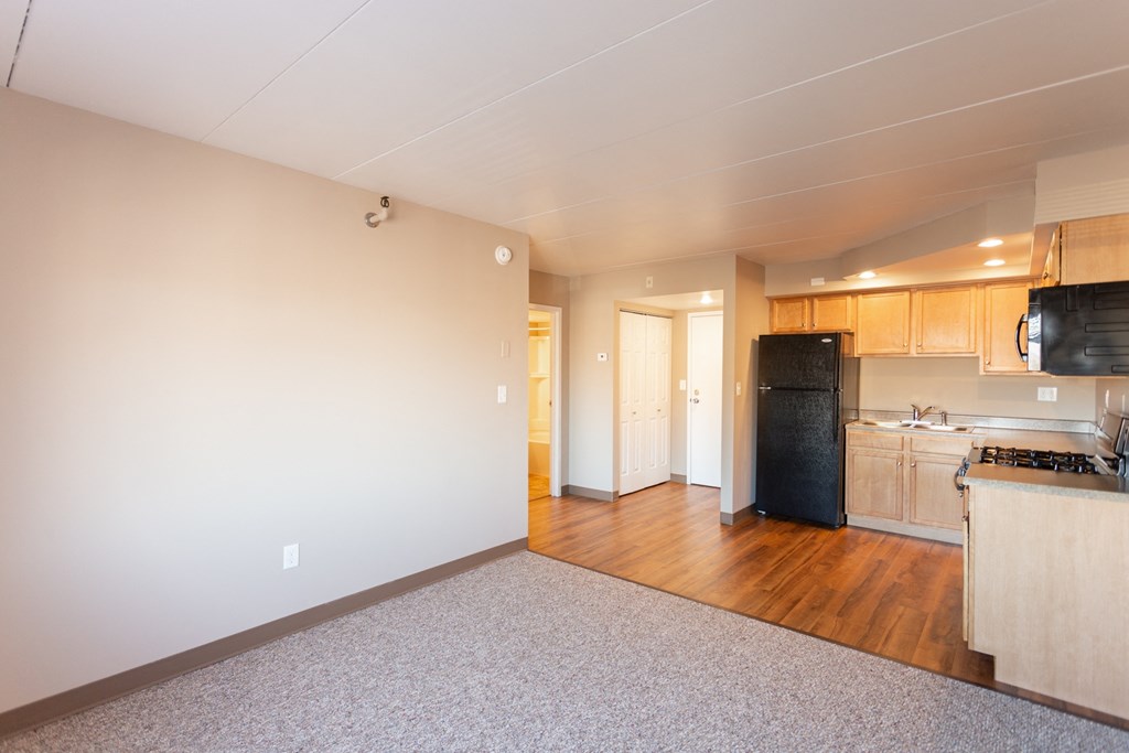an empty living room with a kitchen and a black refrigerator