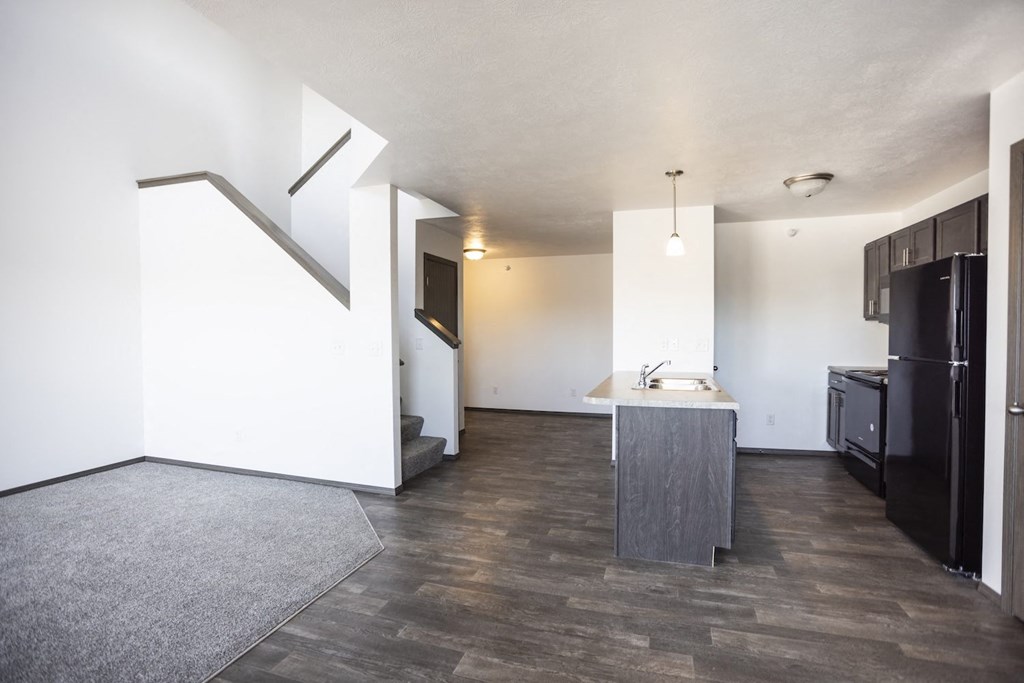 an empty living room with a kitchen and a black refrigerator