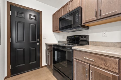 A black door is on the left of a kitchen with a microwave and oven.