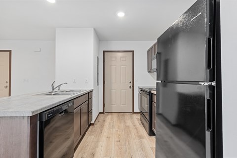 A black refrigerator is in the foreground of a kitchen with wooden floors and white walls.