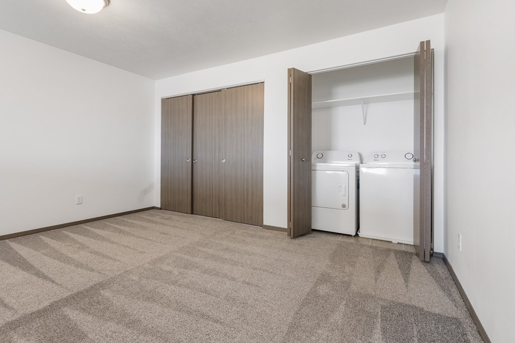 A room with a carpeted floor and a white wall with a fridge and cupboards in it.