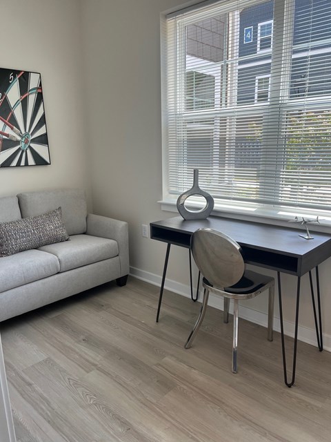 A room with a grey couch, a table with a chair and a dartboard on the wall.