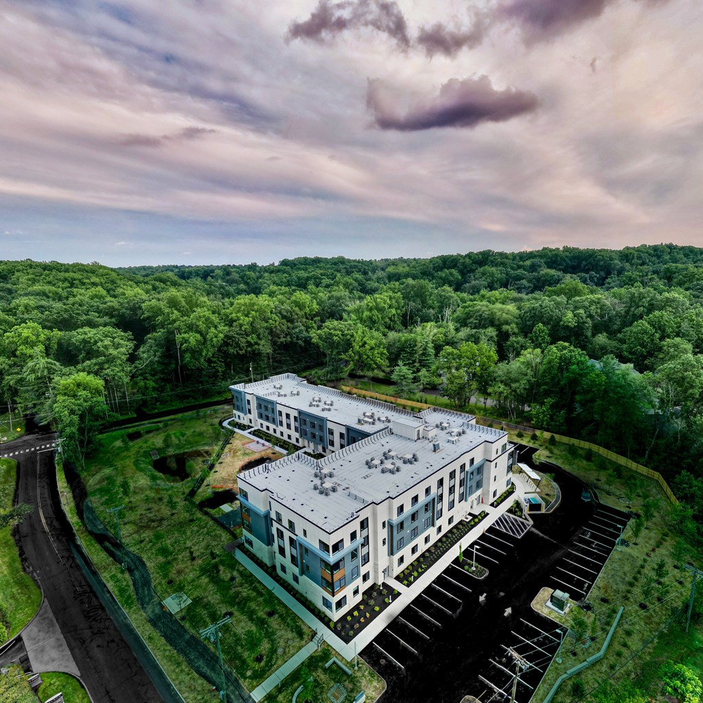 A large white building with a grey roof is surrounded by a parking lot and greenery.
