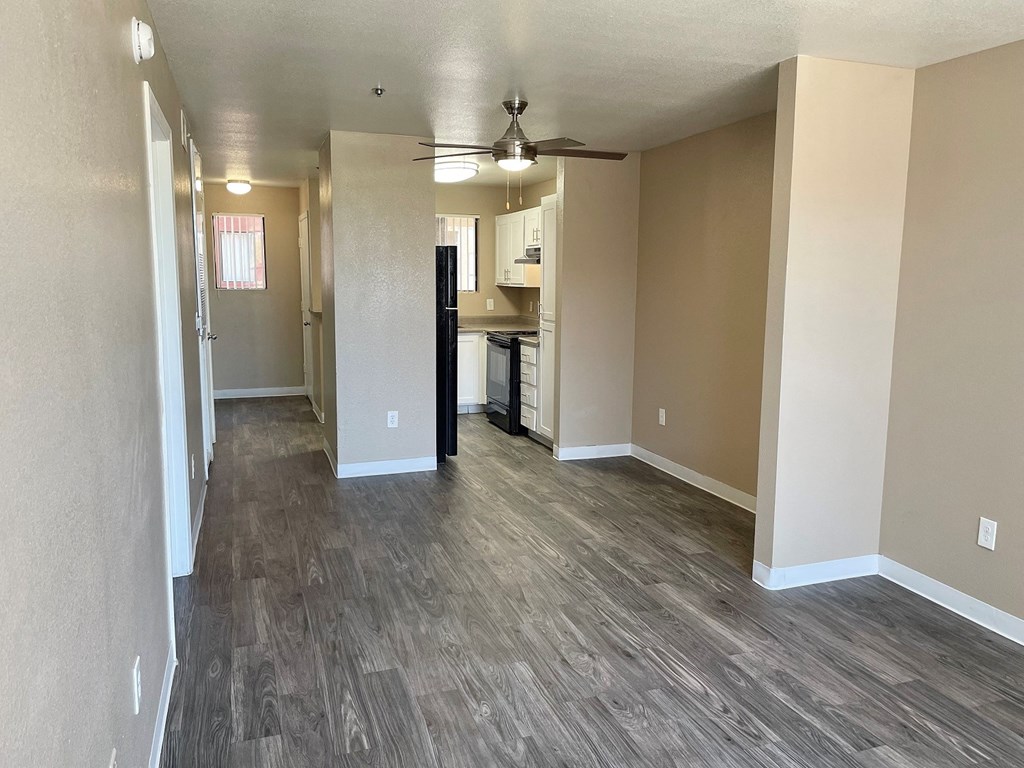 an empty living room with a ceiling fan and a kitchen in the background